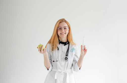 Portrait of a female doctor standing with an apple and a syringe on a light background. The concept of healthy nutrition and medicine.