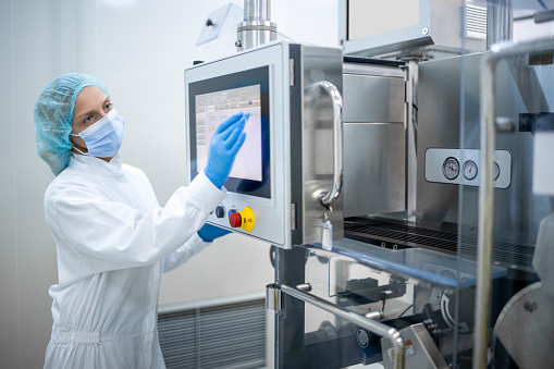 Female employee in a laboratory wearing protective gloves, mask, cap and suit seen in front of the screen of the machine that is the part of the medicaments production during the working hours in a pharmaceutical factory.