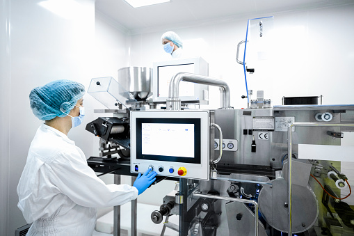 Woman wearing protective gloves, mask, cap and suit seen in front of the screen of the machine that is the part of the drugs production in a laboratory during the working hours in a pharmaceutical factory.