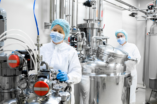 Two women wearing protective gloves, mask, cap and suit seen working on specific machines which are the part of the drugs production in a laboratory during the working hours in a pharmaceutical factory.