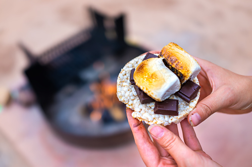Young woman camping closeup hand holding roasted marshmallows smores with chocolate bar squares and rice cake cracker by fire pit in campground campfire grill