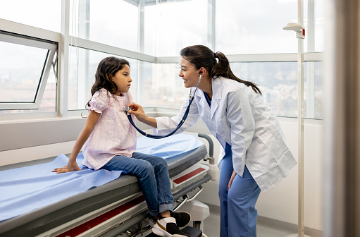 Happy Latin American female doctor examining a little girl at her office and listening to her heart with a stethoscope