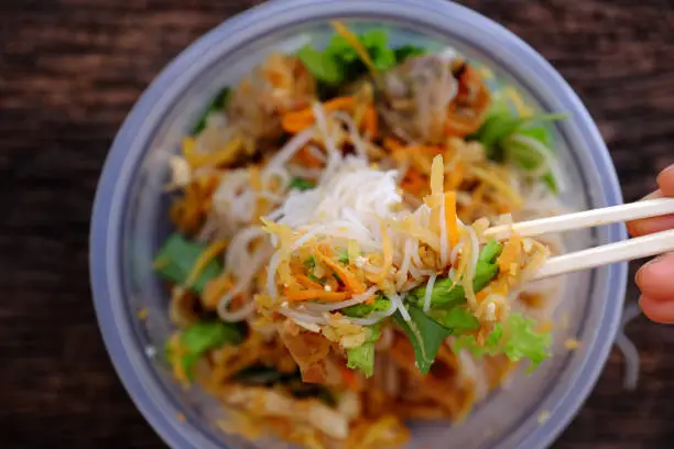 Top view close up hand hold chopsticks pick up food from serving from order online in plastic bowl,  fried spring rolls with vegan rice noodles