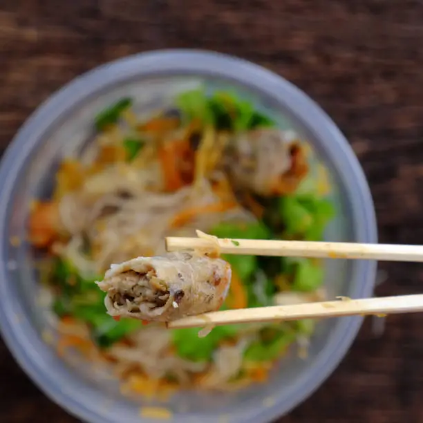 Top view close up hand hold chopsticks pick up fried spring rolls on vegan rice noodles serving from order online in plastic bowl