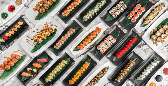 banner with different sets of maki sushi rolls, nigiri and gunkan with tiger shrimp, tomago, salmon, tuna, chuka salad, tuna and bamboo leafs in a white and black plates above a marble table, top view