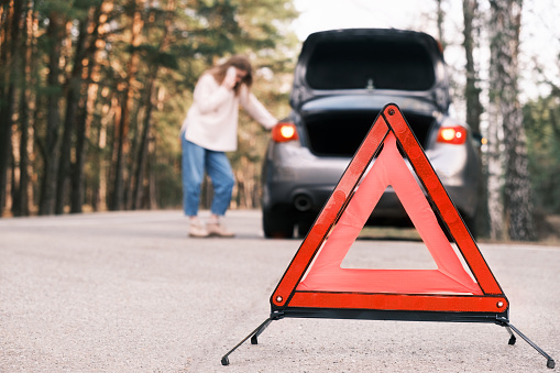 Close up on triangle warning sign with woman calling for assistance after breaking down with her car on background. Broken car on the road.