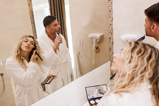 Young couple doing morning routine in bathroom, man shaving beard with electric razor and woman applying face powder