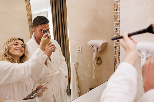 Young couple doing morning routine in bathroom, man shaving beard with electric razor and woman applying face powder