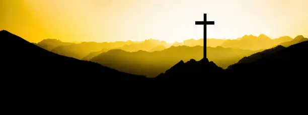 Religious grief landscape background banner panorama - View with black silhouette of mountains, hills, forest and cross / summit cross, in the evening during the sunset, with orange colored sky