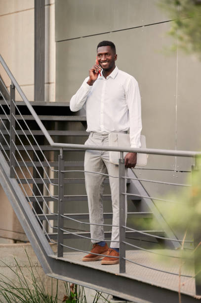 pełne ciało pozytywnego afrykańskiego przedsiębiorcy płci męskiej, który rozmawia przez telefon, stojąc na klatce schodowej w nowoczesnym biurowcu - men black body building african descent zdjęcia i obrazy z banku zdjęć