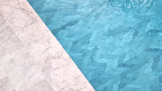 swimming pool background. part of swimming pool showing white carrara marble tile at pool side and blue water with luxury chevron pattern tile under water.
