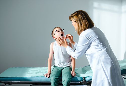 Female doctor checking a sore throat of her young male patient wearing eye glasses. He is sitting on the bed while the doctor is using tongue depressor to have a better look at his throat.
