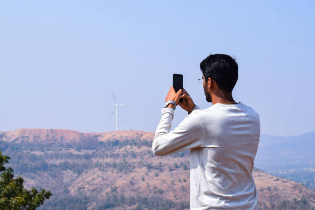 Back view of 25 to 30 age Indian man wearing white color t shirt and spectacles, clicking some photographs from his smart phone of the hills under bright sunlight. stock photo