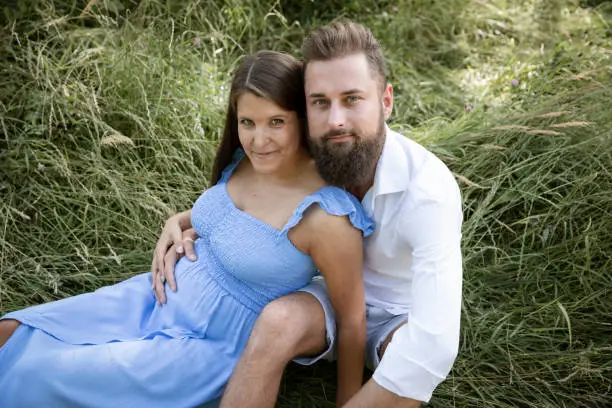 Photo of young couple in love sitting in high flower meadow in summer cuddling and woman is pregnant