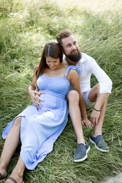 Photo of young couple in love sitting in high flower meadow in summer cuddling and woman is pregnant