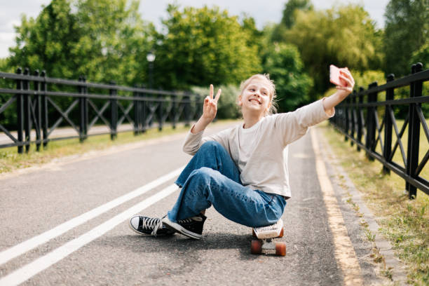 linda adolescente rubia sentada en una patineta en un parque de la ciudad charlando en un teléfono inteligente con amigos y tomándose una selfie. - snowboarding snowboard teenager red fotografías e imágenes de stock