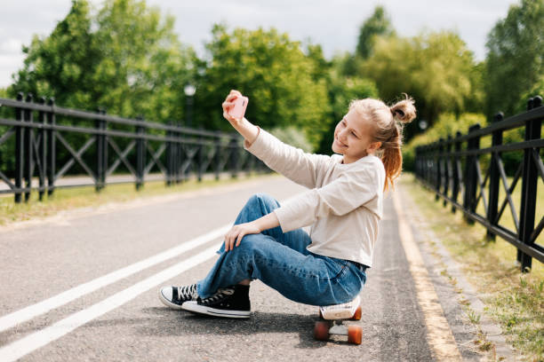 linda adolescente rubia sentada en una patineta en un parque de la ciudad charlando en un teléfono inteligente con amigos y tomándose una selfie. - snowboarding snowboard teenager red fotografías e imágenes de stock