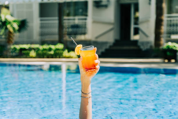 nahaufnahme der hand der frau, die aus dem schwimmbad ragt und einen tropischen cocktail hält. beschnittene mädchenhand mit einer orangenlimonade und dem hotelpool im hintergrund. hallo sommerferien. - drink swimming pool cocktail poolside stock-fotos und bilder