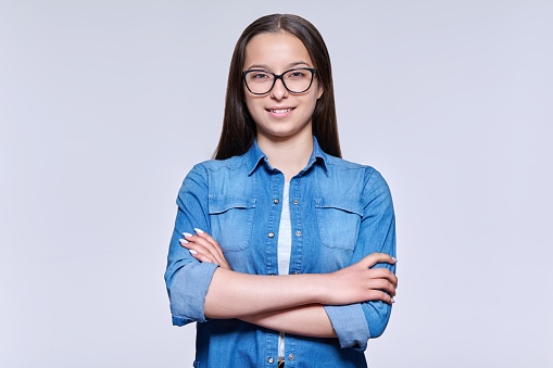 Portrait of smiling beautiful teenage girl in glasses looking at camera on light studio background. Confident female high school student with crossed arms. Education, adolescence, youth, young people