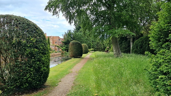 A dirt track path running Parallel to a river by a large stately home with trees and bushes on a sunny day