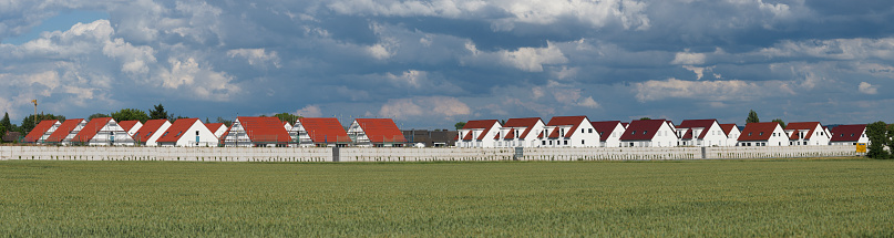 Panoramic view of partially scaffolded semi-detached houses of a new housing estate behind a noise barrier with a green field in the foreground and cloudy sky in the background