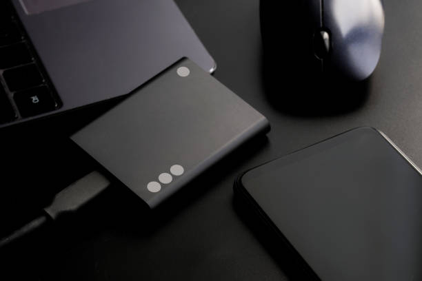 istock Black external portable SSD on black background next to laptop, smartphone and computer mouse. Portable Solid State Drive. Black style. Concept of modern gadgets and information storage 1405067545