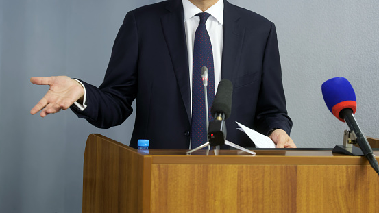 A man - a politician, businessman, official or lawyer gesticulates with his hands, speaking from the podium in front of microphones. Orator, presenter, speaker or interviewee. No face