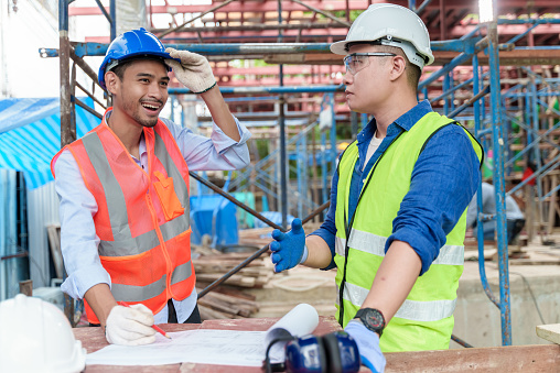 Two young Asian men professional engineer or architect in protective helmet using building design drawing paper checking project progress and standing on worksite. Building engineers working on construction site.