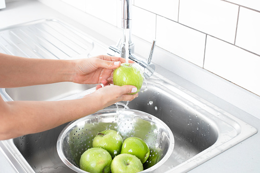 Mid-adult woman rinsing green apples in sink with colander.