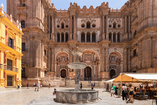 Malaga, Spain, June 10, 2022; Facade of the Malaga Cathedral or Santa Iglesia Catedral Basílica de la Encarnación, with a fountain and tourists in the town square in the foreground.