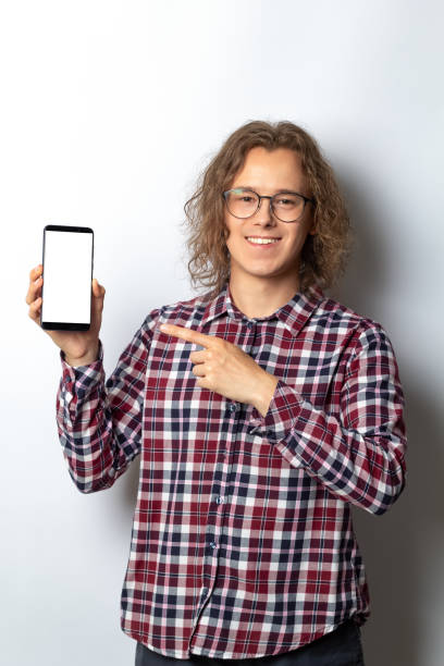 istock Handsome young man holding smartphone with white blank screen. The guy points his finger at the blank mobile phone mockup. 1405104964
