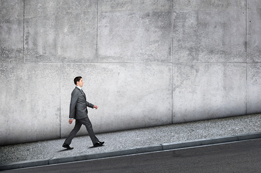 A businessman walks uphill on a sidewalk next to a large concrete wall that provides ample room for copy and text.