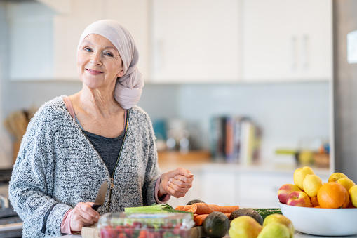 A woman wearing a headscarf stands by the kitchen counter, which has a colourful array of vegetables on it. She smiles as she chops the produce.