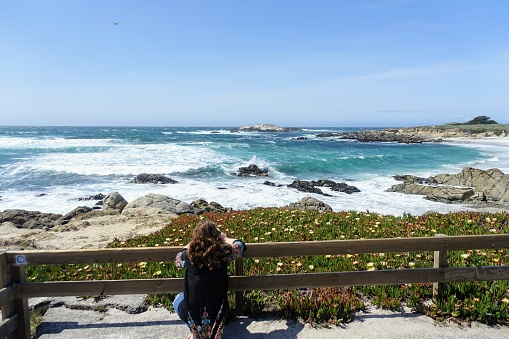 A woman admiring the beautiful coastal views along the 17 mile drive outside Carmel by the Sea, which bright blue ocean scenery and waves crashing against the rocky shore