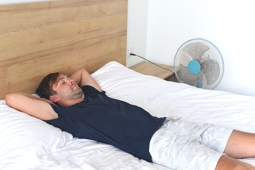 Young man laying and sleeping against fan on a hot summer day