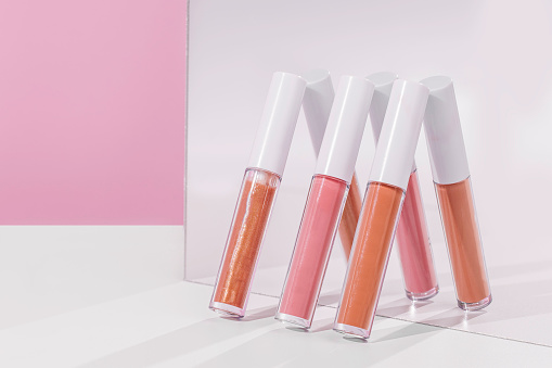 Three different shades of lip gloss are leaning up against a mirror. There is a pink background and a white table.