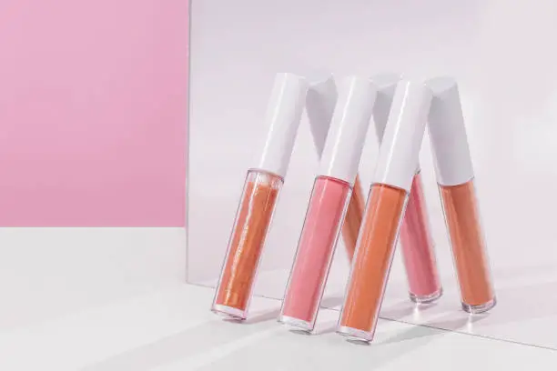 Three different shades of lip gloss are leaning up against a mirror. There is a pink background and a white table.