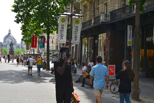 Antwerp city, Province Antwerp, Belgium - June 18, 2022: African man using smart phone. Tourists, citizens walking the street Leys. Antwerp station on the horizon. Veritas DIY sewing and thread shop