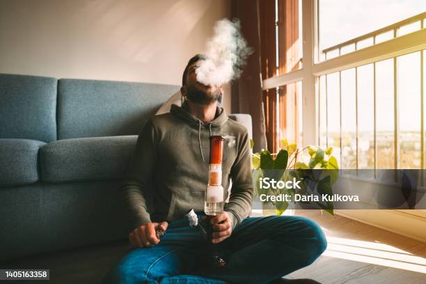 Man Smoking Bong And Exhaling The Smoke At Home Man Smoking Pot Medical Marijuana Or Cannabis From A Bong Or Water Pipe Cannabis And Weed Legalisation Concept Stock Photo - Download Image Now