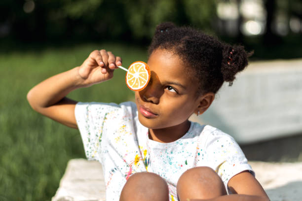 A cute African-American girl covers one eye with a bright fruit lollipop in the park on a summer day.Summer holidays,diverse people concept