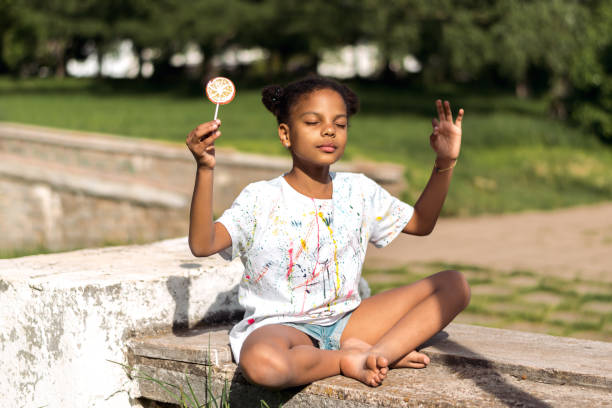 A cute African-American girl with lollipop in her hands sitting in the lotus position on a summer day in the park  and enjoying the sun.Summer concept,diverse people concept