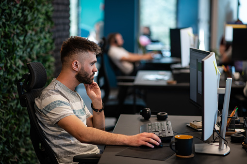 Young IT expert working in his modern and new coworking office. Programmer typing at his work place, coding on his computer. Man sitting at his desk in a joint office, with colleagues in the background.