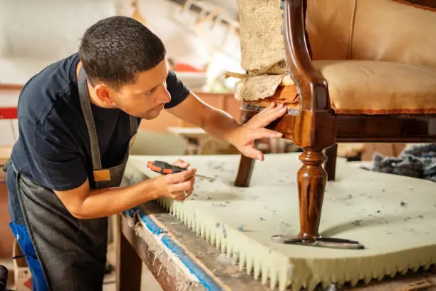 Upholstery worker attaching new batting to an arm chair sitting on a bench in a furniture workshop