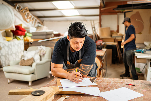 Worker sketching design ideas on some paper while leaning on a workbench in furniture making shop