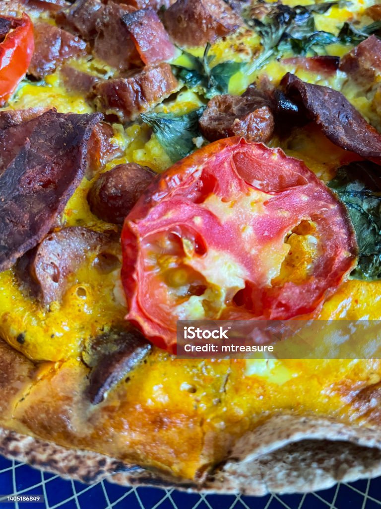 Full frame image of Spanish omelette frittata on blue rimmed plate, pepperoni slices, sausage, cherry tomato halves, wilted spinach leaves, elevated view, focus on foreground Stock photo showing close-up, elevated view of frittata egg omelette containing pepperoni slices, sausages, cherry tomato halves and spinach leaves. Breakfast Stock Photo