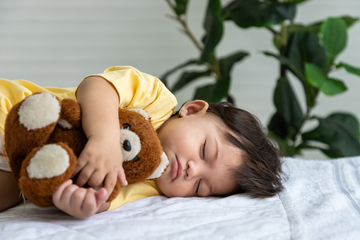 Little Asian baby girl lying down sleeping on bed at home, hugging teddy bear doll. Newborn child relaxing in bed. Nursery for young children.