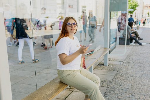 A cheerful caucasian woman is sitting on a bench inside of an urban bus stop and using her cell phone and paper map. Urban travel and transportation concept. Girl search for the location