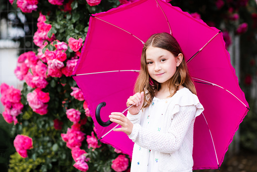 Smiling child girl 8-9 year old hold umbrella standing at city street in park over rose flowers outdoor close up. Portrait of happy blonde child over nature background. Summer rain.