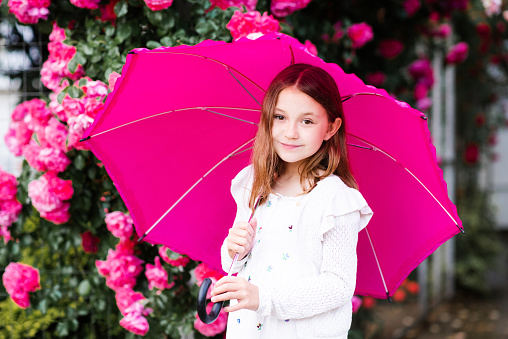 Smiling child girl 8-9 year old hold umbrella standing at city street in park over rose flowers outdoor close up. Portrait of happy blonde child over nature background. Summer rain.