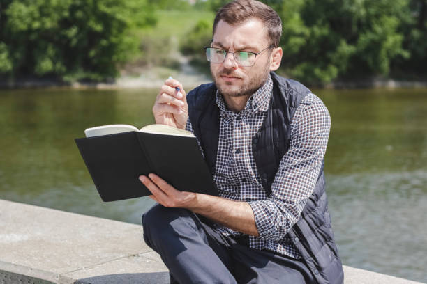 istock Intelligent man wearing glasses looking at his diary and writing notes while sitting outdoors near the river. 1405213124